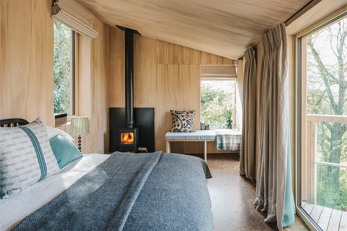 Elmore Treehouses bedroom interior with wood-burning stove and plywood walls overlooking woodland